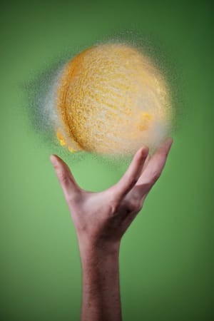Photo of a burst water balloon. It’s light yellow, and held in a hand.