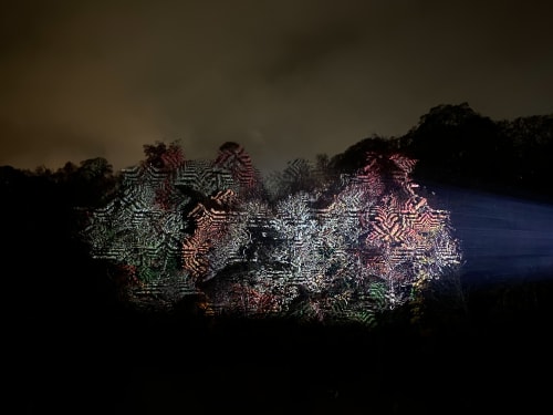 A projector projects repeating patterns on to the trees on a far riverbank.
