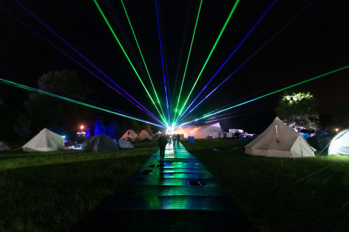 Looking straight down a path at night with white tents on either side. In the distance are lasers starting at the end of the path and going out in all directions in the sky.