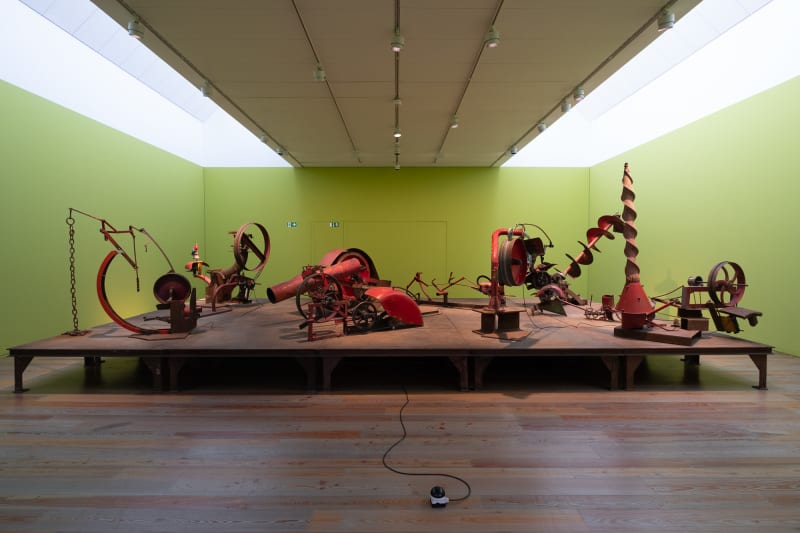 Wide photo of pale green room with several red mechanical sculptures in the middle.