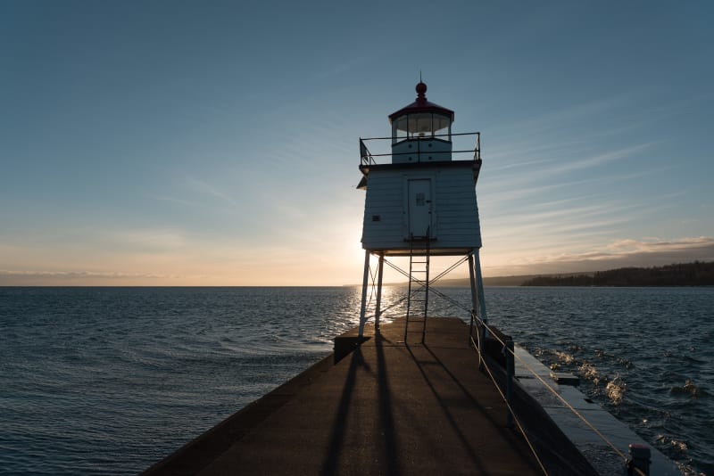 A small lighthouse at the end of a pier is silhouette in front of a setting sun.