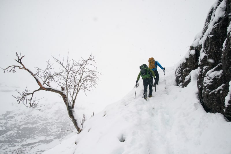 Two hikers climb a somewhat steep snowy hillside. It’s overcast with flurries of snow around.