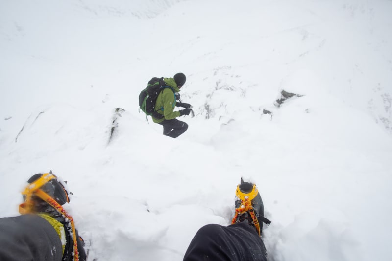 Two boots with crampons on are in the foreground descending a steep snowy mountain. Ed can be seen a few meters ahead.
