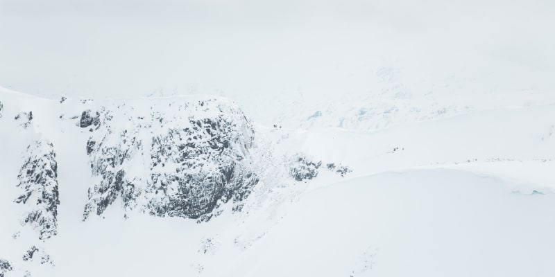 A long telephoto photo of a snowy ridge in the distance. A few small brightly coloured figures can be seen on the ridge.