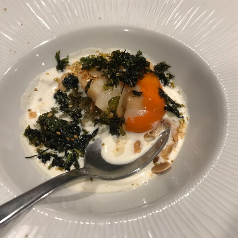 A close up of a bowl of food. The bowl contains a single scallop in a white cream sauce. There’s scattered fried seaweed on top.