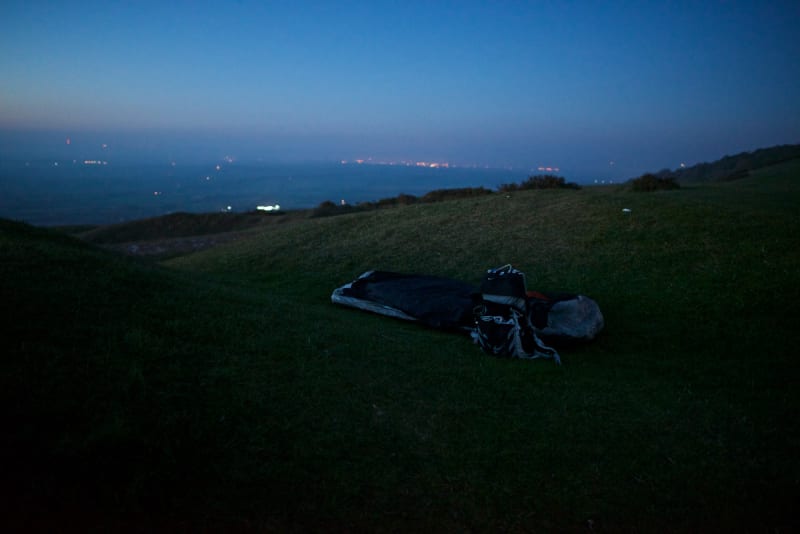 A bivvy bag and rucksack sit in a hollow depression in the grass in the last of the evening light. The sky is dark blue and calm.