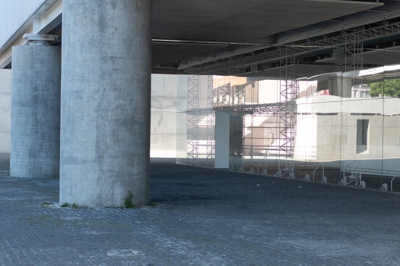 A photo of an exterior walkway near the National coach museum. On the right is a glass exterior wall of the building with strong reflections obscuring the inside. On the left are two wide concrete columns supporting the walkway.