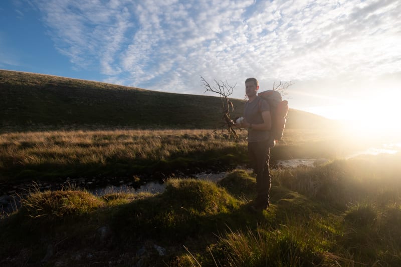 A hiker stands still looking at the camera holding bunches of sticks in each hand. He’s lit from behind by golden sunlight.
