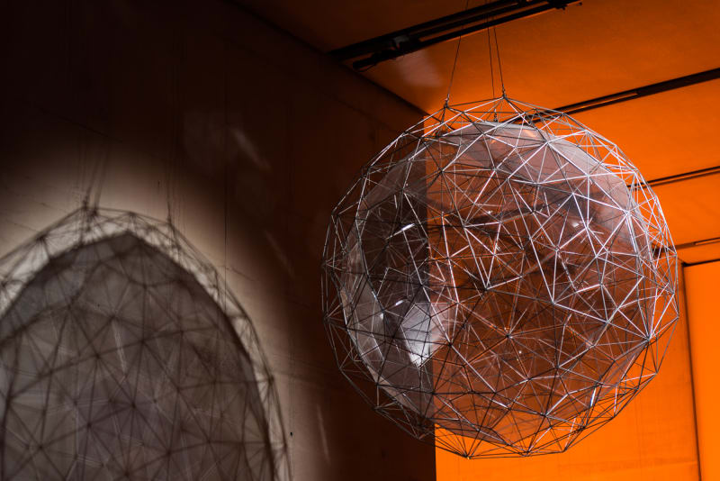 A semi-solid sphere of made from metal rods hangs against an orange backdrop.