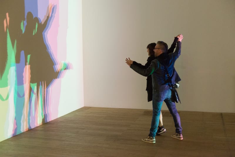 Two people hold hands in a room. A light shines on them from one side, casting a multi-coloured shadow of them against the wall.