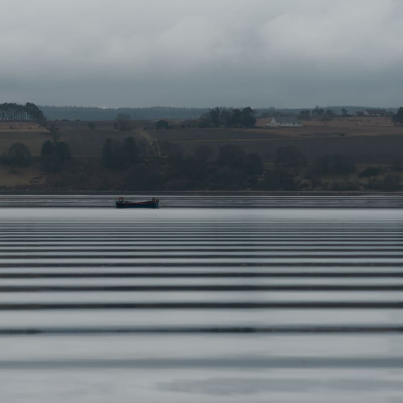 Looking across a wide lake on a grey day. In the distance is a small industrial boat. The water has regular horizontal dark bands - like a barcode - caused by ripples from a passing boat.