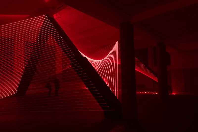 Red lasers in a dark room form lines that sweep up to a column then down again like threads.