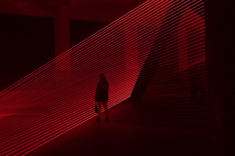 Red lasers in a dark room form even horizontal lines going from bottom left to upper right. A figure is silhouetted in front of them.