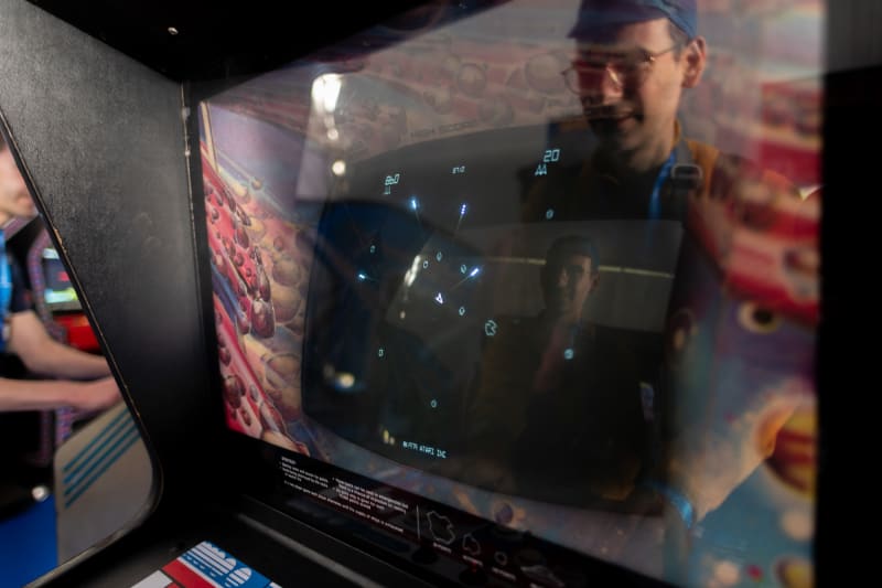 Close up photo of the screen of a vintage Asteroids arcade game with dark black screen and white ship and asteroids. There’s strong reflections on the cover glass and the player’s face is reflected twice.