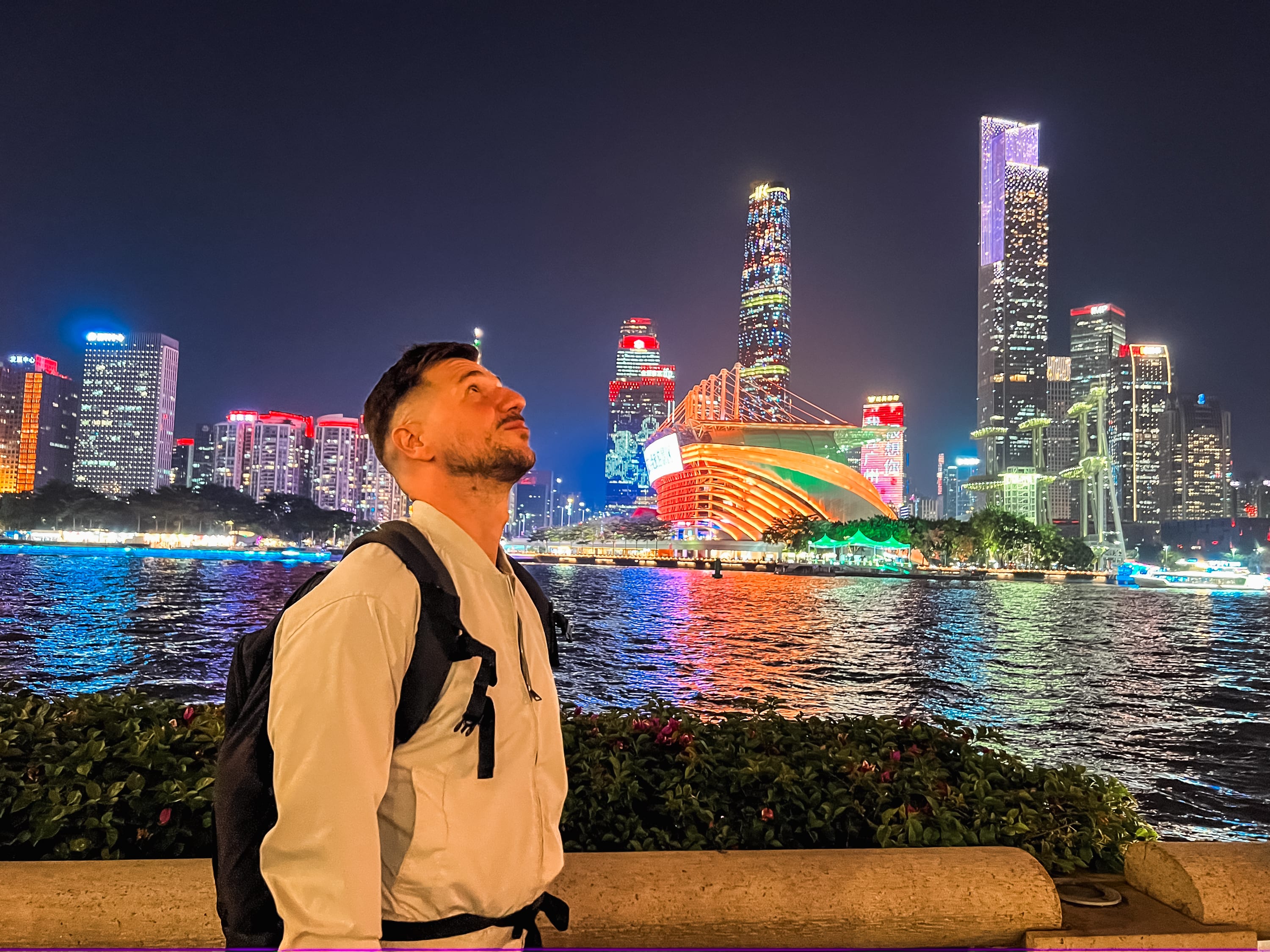 A man with a backpack stands by a waterfront at night, looking up. The vibrant urban skyline of Guangzhou glows with bright lights, reflecting the colorful energy of gay life in China across the water.