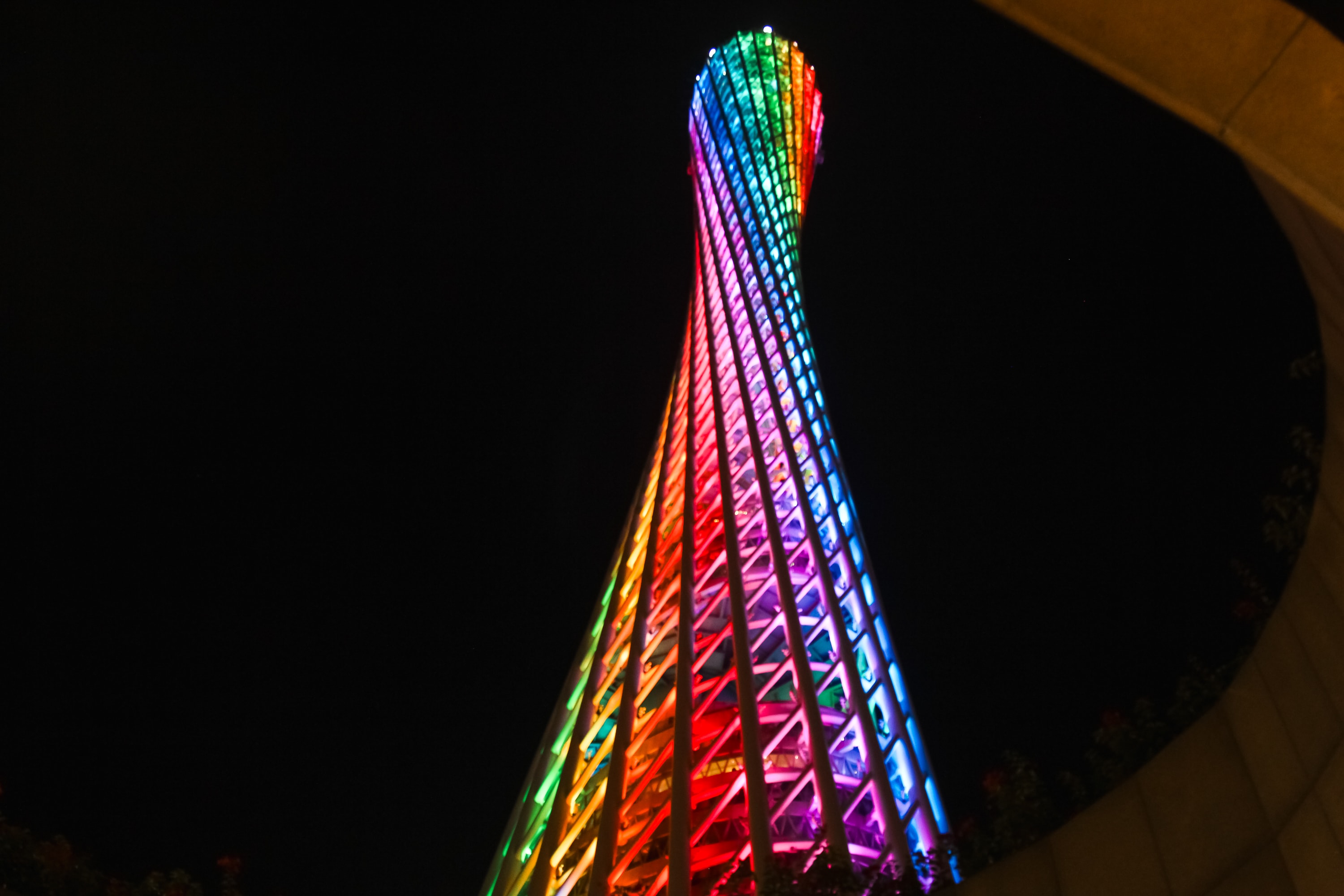 A tall, twisting tower illuminated with vibrant rainbow-colored lights stands against a dark night sky in gay Guangzhou, viewed from a low angle.