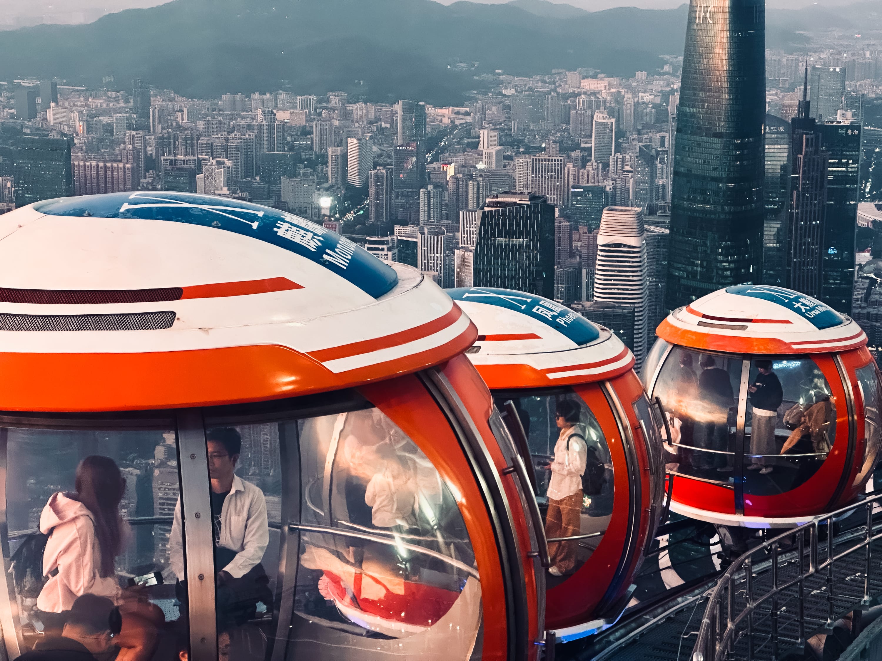 People sit inside colorful glass observation pods on a rooftop track in gay Guangzhou, overlooking a sprawling city skyline with tall buildings and distant mountains at sunset.