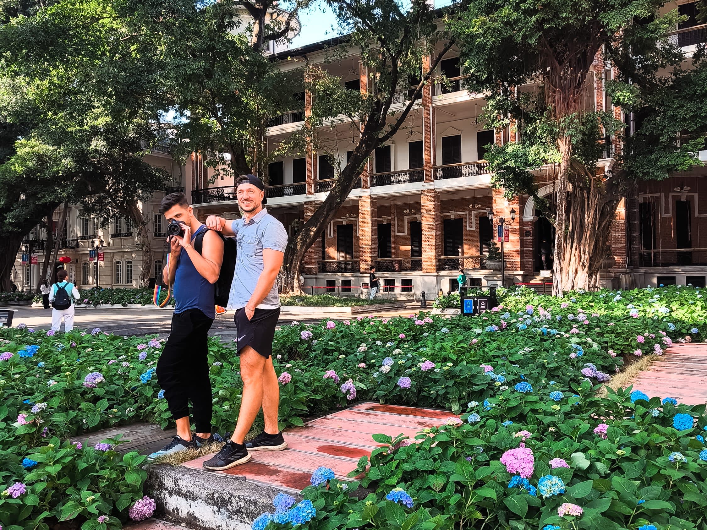 Two men stand smiling among blooming hydrangeas in front of a historic brick building in Guangzhou. Celebrating gay life in China, one holds a camera while the other looks on, both appearing relaxed and happy amid large trees and balconies.