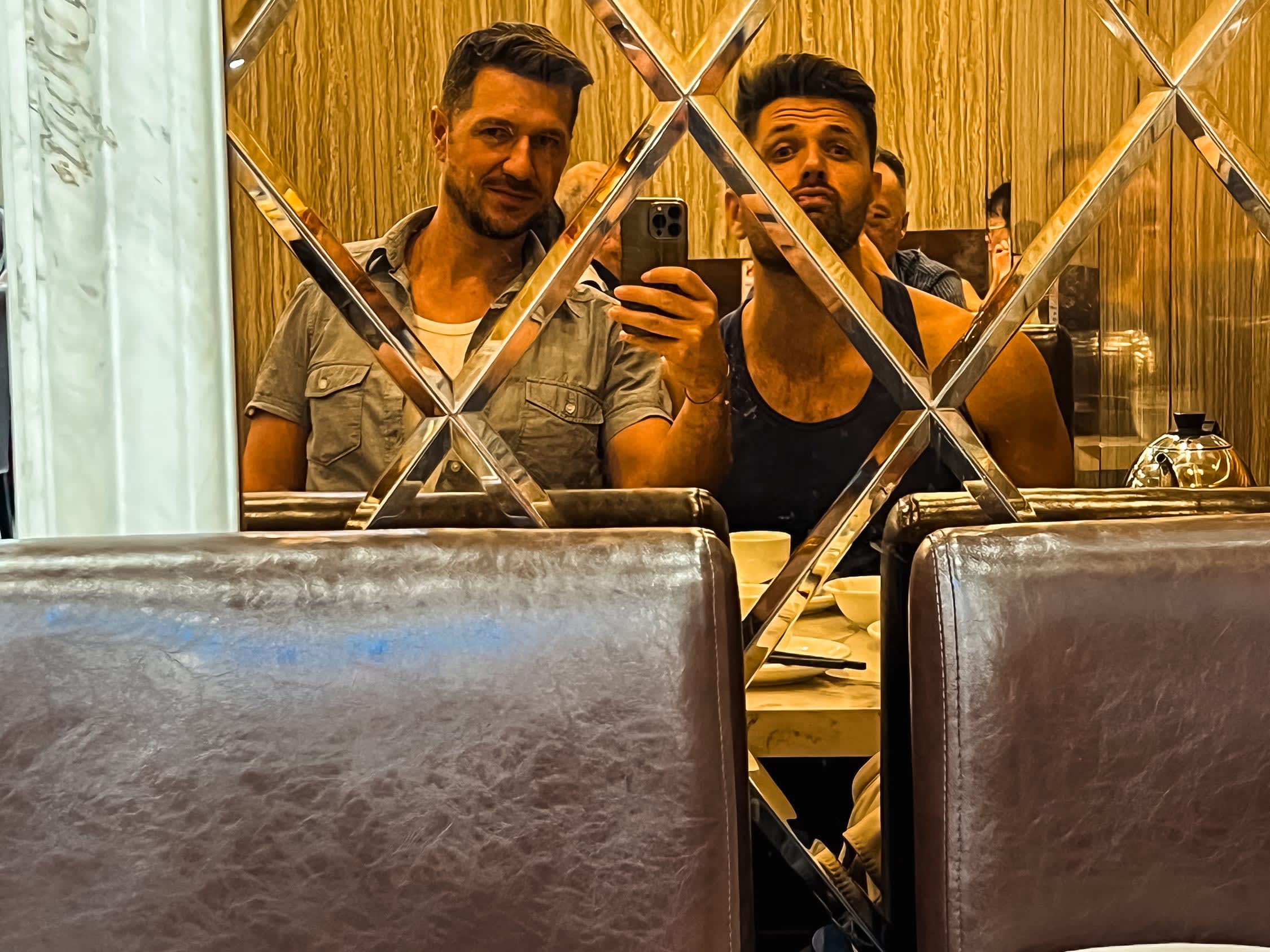 Two men sit side by side in a restaurant, taking a mirror selfie. Reflected in a diamond-patterned mirrored wall, they capture a candid moment of gay life China, with cups and plates visible on the table before them.
