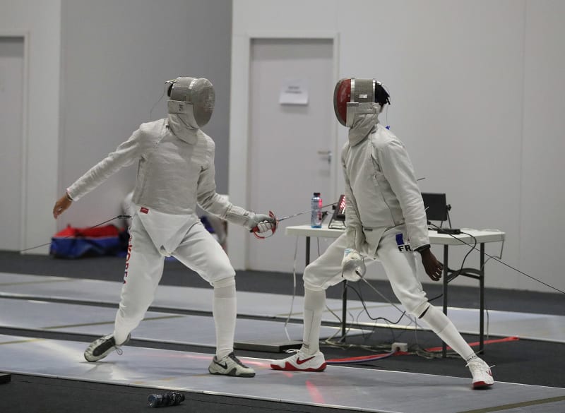 Dos esgrimistas en un asalto de entrenamiento sobre la pista, ambos con careta; de sus espaldas salen los cables de cuerpo hacia el aparato de señalización en una mesa al borde de la imagen