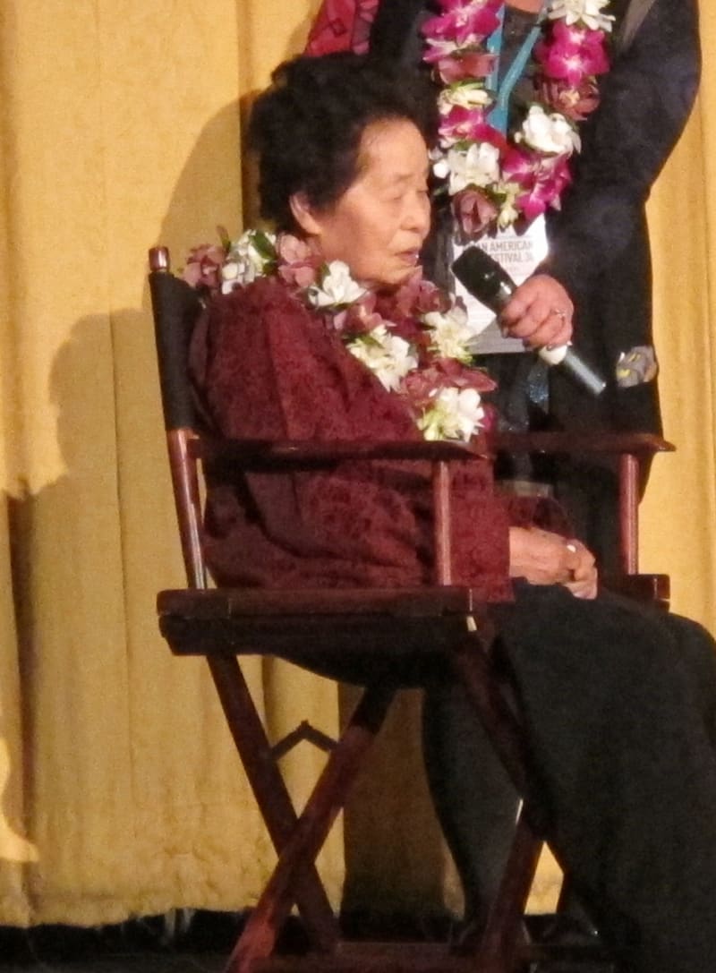 Keiko Fukuda mit 98 Jahren auf einer Bühne in San Francisco, März 2012: sie sitzt in einem Regiestuhl, trägt eine Blumenkette und antwortet, während jemand neben ihr das Mikrofon hält