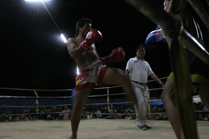 Kun-Khmer-Kampf in Battambang, Kambodscha, 2006: Ein Kämpfer in roter Hose und Boxhandschuhen hebt das Knie, dahinter ein Ringrichter in Weiß; ringsum drängt sich das Publikum am Seilring