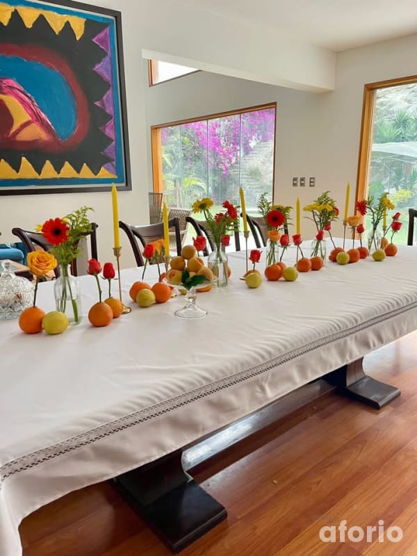 Comedor interior con mesa larga adornada con frutas, velas amarillas y rosas rojas, frente a ventanal con vista al jardín