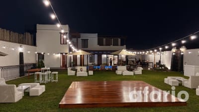 Jardín de noche con pista de baile de madera, muebles lounge blancos y guirnaldas de luces frente a la casa iluminada