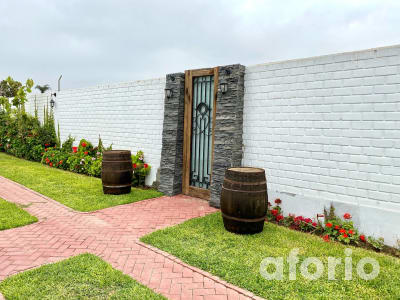 Ingreso al jardín con puerta de reja y marco de madera sobre muro blanco, barricas de vino decorativas, camino de ladrillo y jardineras con geranios rojos