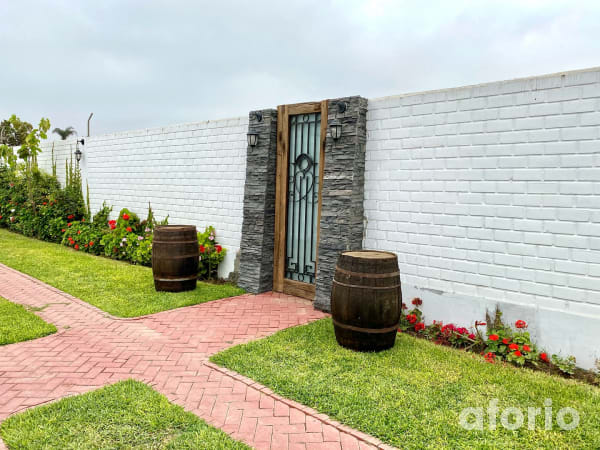 Ingreso al jardín con puerta de reja y marco de madera sobre muro blanco, barricas de vino decorativas, camino de ladrillo y jardineras con geranios rojos