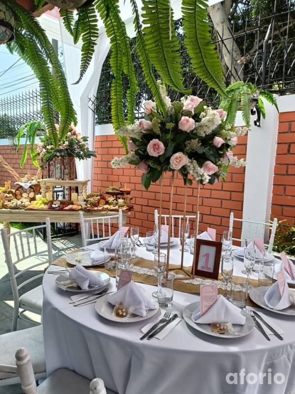 Mesa redonda montada para evento con mantel blanco, sillas Tiffany, centro alto de rosas rosadas y mesa de bocaditos al fondo
