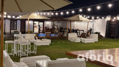 Zona lounge nocturna junto a la piscina con sombrillas, mesas altas con banquetas blancas y terraza techada al fondo