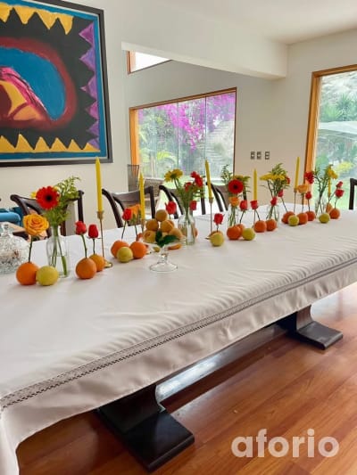 Comedor interior con mesa larga adornada con frutas, velas amarillas y rosas rojas, frente a ventanal con vista al jardín