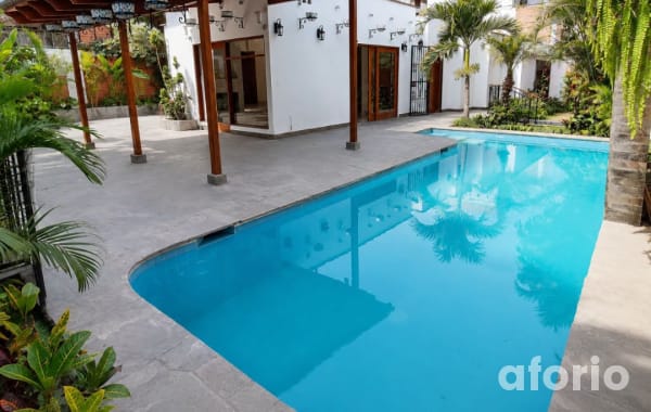 Piscina de agua turquesa bordeada de palmeras, con terraza de piedra y pérgola de madera junto a la casa blanca
