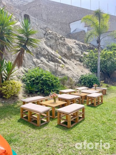 Zona lounge en el jardín con mesas y banquetas de madera sobre el césped, con vista al cerro