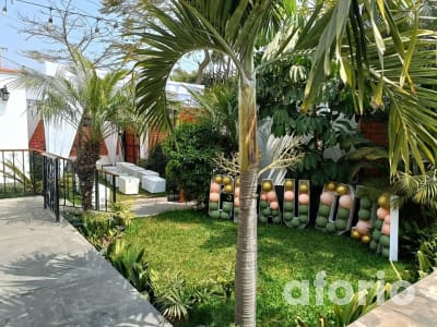 Jardín con palmeras y letras decorativas de globos verdes y rosados, junto a muebles lounge blancos bajo carpa