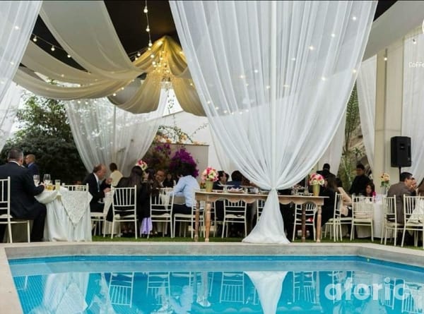 Recepción de evento bajo telas blancas drapeadas con araña de cristal, mesas con sillas Tiffany y piscina al frente
