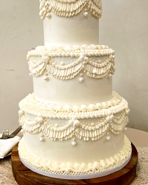 Wedding cake by Sweet E's Bakery in Mapleton, a three-tier, round, smooth buttercream cake in ivory, adorned with piped swags, rosettes, and