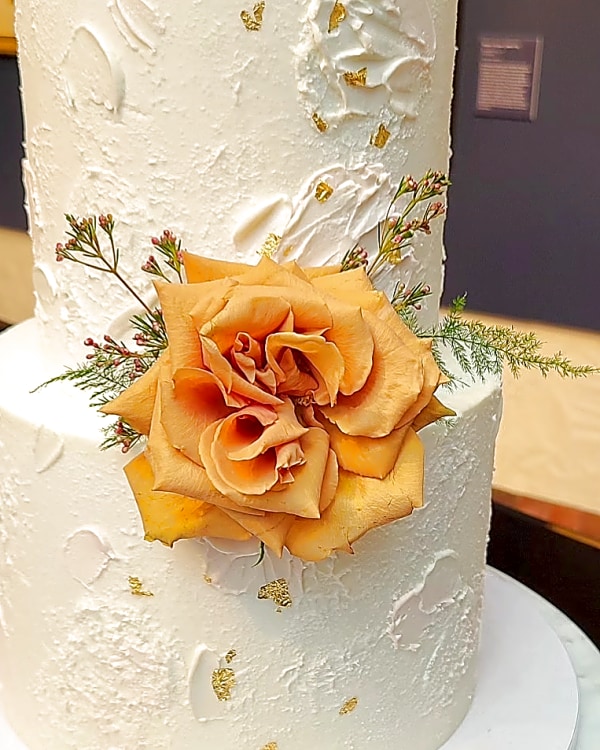 Wedding cake by Sweet E's Bakery in Springville, two-tier white cake with textured finish, gold leaf accents, and a large orange rose with small