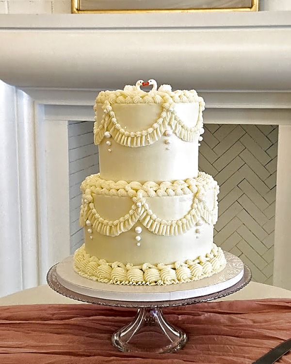 Wedding cake by Sweet E's Bakery in Spanish Fork, two-tier ivory buttercream cake with intricate piped swags, pearl details, and a ruffled base
