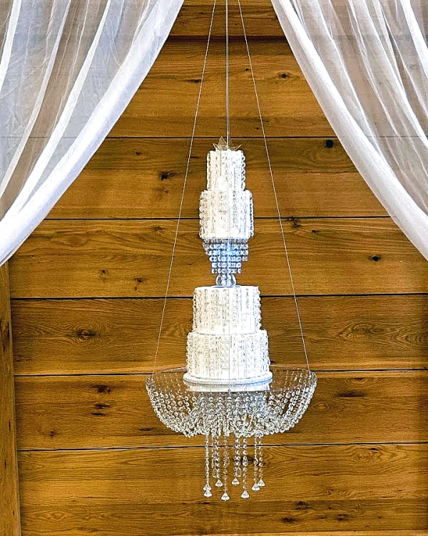 Suspended white textured wedding cake hung from crystal chandeliers against a timber wall