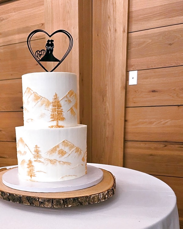 Snowpine Lodge wedding cake, two-tier white buttercream cake with gold painted mountains and trees, topped with a heart-shaped