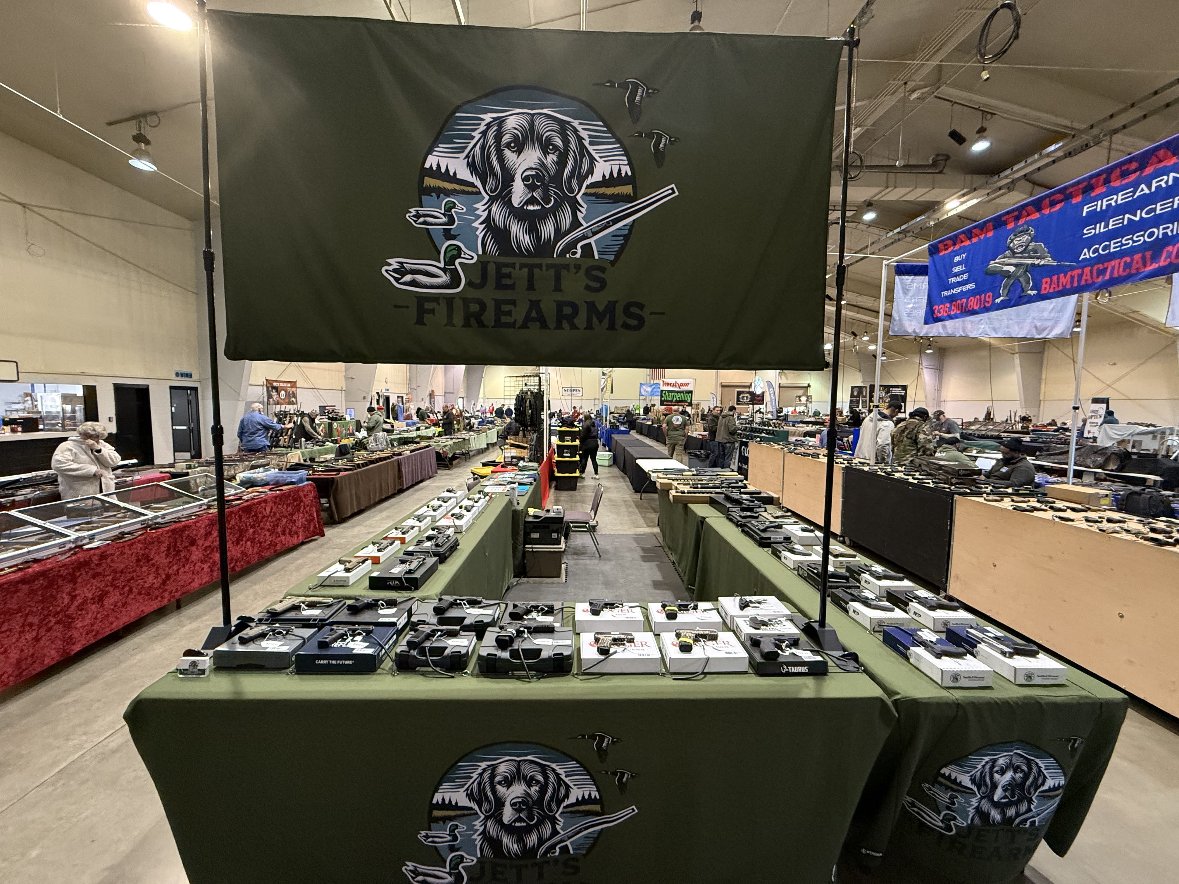 Jett's Firearms gun show vendor table in North Carolina — green branded tablecloth and banner with handguns, rifles, and firearms on display