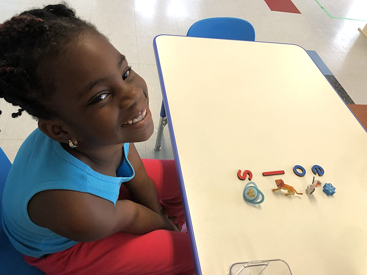 Garderie Ami Câlin : une enfant sourit à la caméra, concentrée et confiante, lors d'un atelier langage Montessori avec lettres et petits objets sur une table d'éveil. — Garderie Ami Câlin — Instantané d'apprentissage joyeux — Un moment de bonheur à apprendre en environnement Montessori. Inscriptions à la petite enfance en cours — garde et éducation professionnelles.
