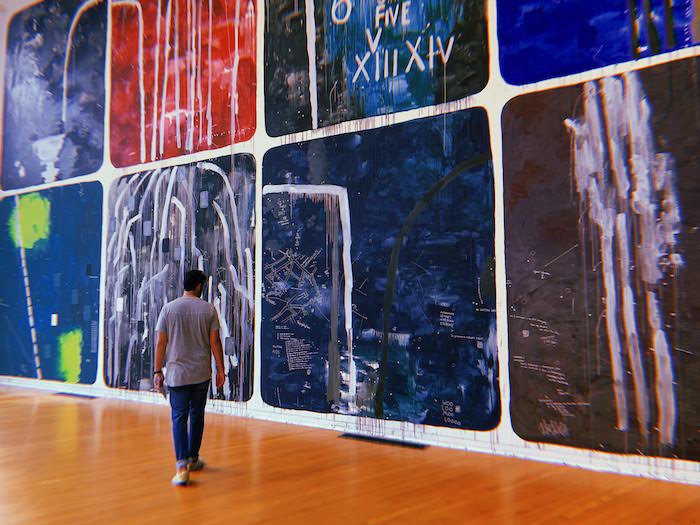 man with dark hair walking in front of a large indoor art mural