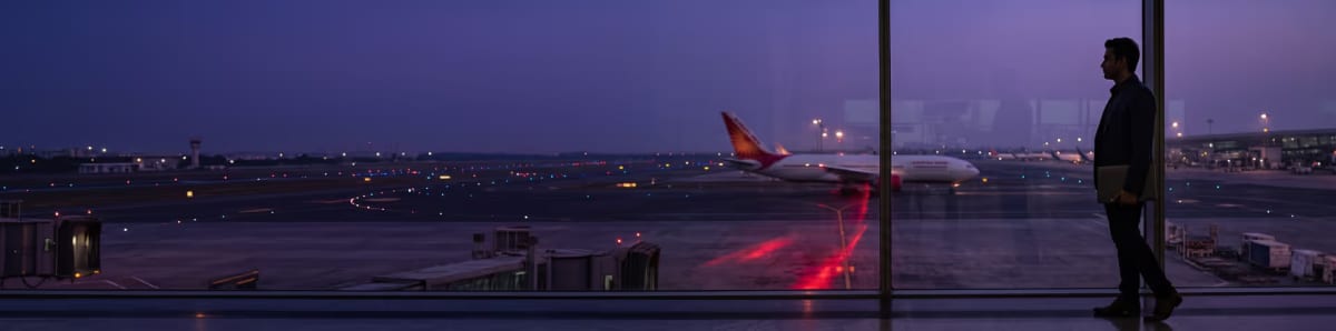 A traveller with a laptop stands at an airport terminal window at dusk, watching an airliner on the apron