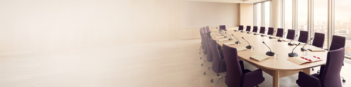 An empty council chamber with a long oval conference table, purple chairs, a microphone and a folder at every seat, and floor-to-ceiling windows overlooking a city