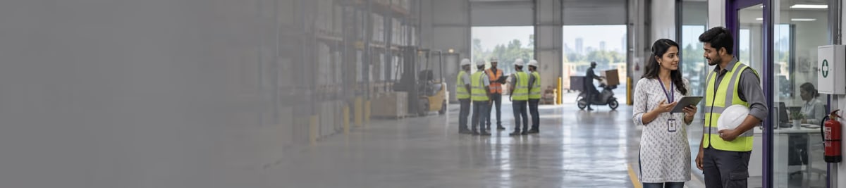 A compliance officer holding a tablet talking with a supervisor carrying a hard hat at a warehouse doorway, a group of workers in high-visibility vests further down the bay