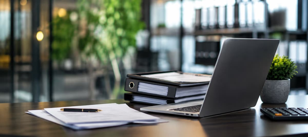 An open laptop, ring binders of paperwork and a signed document on a dark office desk.