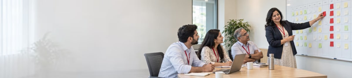 A colleague placing a red sticky note on a whiteboard calendar of colour-coded notes while three seated teammates look on from a meeting table